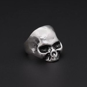 May include: A silver-colored skull ring with a textured surface. The ring features a detailed skull design with hollow eye sockets and a defined nose and mouth. The ring is a gothic or biker-style accessory.