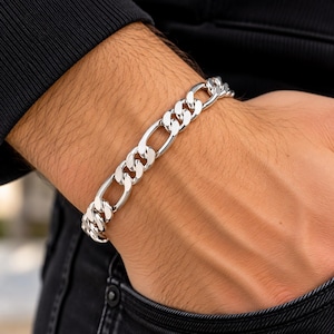 May include: A silver-toned Figaro chain bracelet featuring a repeating pattern of three small circular links followed by one elongated oval link. The metal surface is polished and reflective, creating a smooth, continuous metallic appearance.