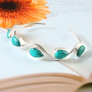 Peut inclure: Un bracelet manchette en argent orné de cinq pierres turquoise en forme de goutte. Le bracelet présente un design ondulé. Fond blanc avec une fleur orange en arrière-plan.