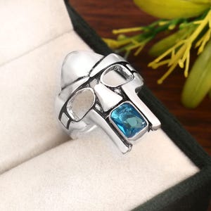 May include: A silver-colored ring with a unique, angular design. The ring features a rectangular, light blue gemstone set in the center. The ring's design is reminiscent of a mask or helmet.