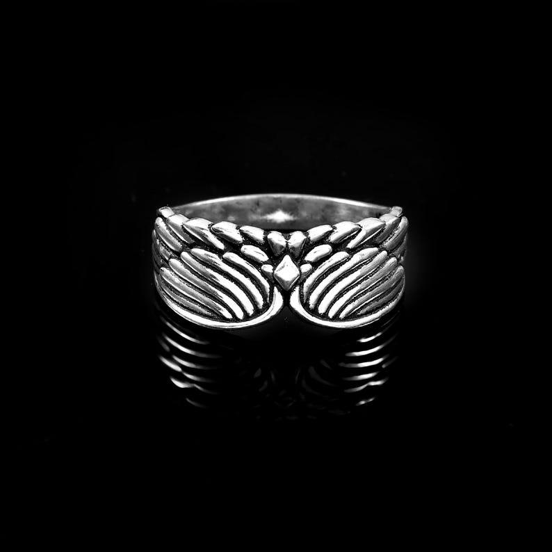 May include: A silver-toned metal ring featuring a symmetrical wing design that wraps around the band. The surface of the ring has a polished finish with darkened recessed areas to highlight the detailed feather textures and a central diamond shape.