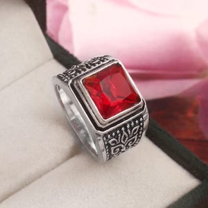 May include: A silver-toned ring with a large, square, red gemstone. The ring has an ornate, engraved design around the gemstone and on the band. The ring is displayed in a dark green ring box.