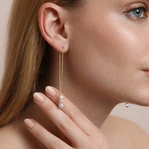 以下が含まれることがあります： 下部に3つの小さな丸い透明な石がぶら下がった、金のチェーンイヤリングです。