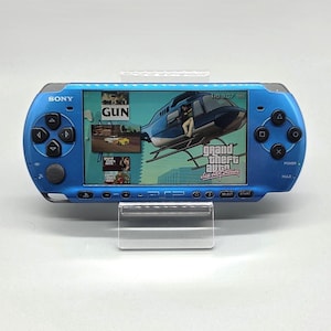 Może przedstawiać: Jasnoniebieska przenośna konsola do gier Sony PlayStation Portable z szerokim ekranem wyświetlającym menu gry Grand Theft Auto Vice City Stories z grafiką helikoptera i napisem GUN oraz różnymi przyciskami nawigacyjnymi.