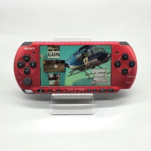 Pode incluir: Uma consola de jogos portátil Sony PSP vermelha com um ecrã panorâmico que apresenta a interface do jogo Grand Theft Auto Vice City Stories, incluindo um gráfico de helicóptero, o texto GUN e vários ícones de menu na caixa de plástico.