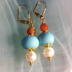 Op de afbeelding: Een paar oorhangers met drie kralen per oorhanger. De bovenste kraal is een kleine, oranje-bruine carneol. De middelste kraal is een grotere, lichtblauwe kraal. De onderste kraal is een witte parel.