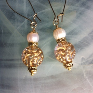 May include: A pair of gold earrings with a white pearl and a gold filigree bead. The earrings have a simple hook closure.
