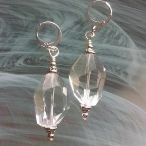 May include: A pair of clear, faceted crystal earrings with silver-colored metal accents. The earrings have a geometric, elongated shape and are suspended from lever-back closures. The crystals catch the light, creating a sparkling effect.
