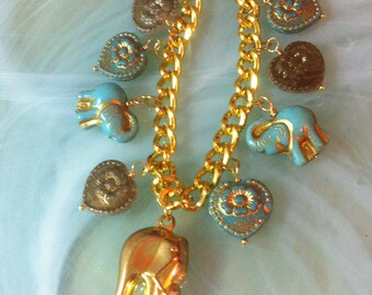 May include: Gold-toned charm bracelet featuring a series of decorative charms. The bracelet includes elephant, heart, and circular charms in gold, turquoise, and brown. The bracelet is made of a gold chain and has a lobster clasp.