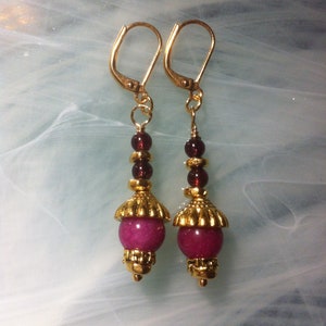 Peut inclure: Une paire de boucles d'oreilles pendantes dorées avec des perles roses et bordeaux. Les boucles d'oreilles ont une fermeture à crochet dorée et une pierre rose au bas.