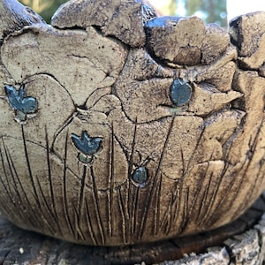 Könnte beinhalten: Eine handgefertigte Keramikschale mit einer strukturierten, erdigen Optik. Die Schale hat ein geprägtes Design aus stilisiertem Gras und kleinen, dunkelblauen Insekten- und Blumendetails. Die Schale steht auf einer verwitterten Holzoberfläche.