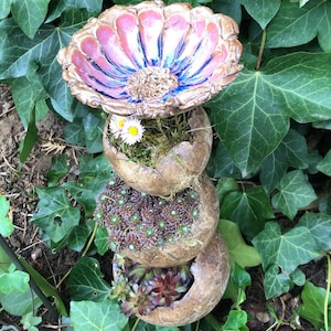 Könnte beinhalten: Ein Vogelbad aus Keramik mit einem pinkfarbenen und blauen Blumendesign, gestapelt auf drei braunen Keramikbehältern mit grünen Sukkulenten und weißen Gänseblümchen.