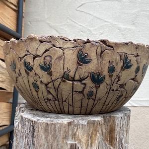 Könnte beinhalten: Handgefertigte, beige Keramikschale mit strukturierter Oberfläche und floralem Design. Die Schale hat dunkelblaue Blumen- und Stieldetails. Die Schale steht auf einem Holzstumpf.