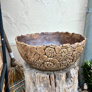 Könnte beinhalten: Handgefertigte, braune Keramikschale mit strukturierter Oberfläche und fossilartigen Mustern. Die Schale steht auf einem verwitterten Holzstumpf und zeigt ihr einzigartiges, organisches Design. Der Rand der Schale hat eine raue, unebene Kante.