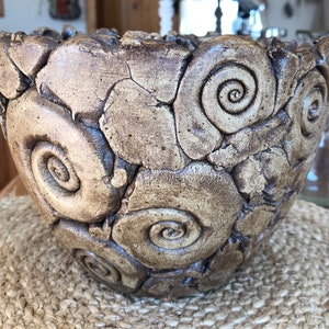 Könnte beinhalten: Eine handgefertigte Keramikschale mit einem einzigartigen Design. Die Schale weist eine Reihe von Spiralmustern und eine rissige Oberfläche auf, wodurch eine rustikale Ästhetik entsteht. Die Farbe ist eine Mischung aus Braun und Beige, was ihren erdigen Reiz verstärkt.