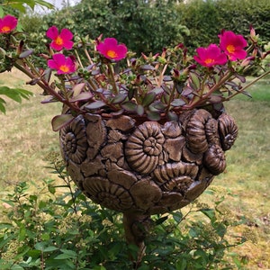 Könnte beinhalten: Ein bronzefarbener Blumentopf mit Fossilienmotiven, gefüllt mit leuchtend rosa Blüten mit gelben Zentren. Der Topf hat eine detaillierte, strukturierte Oberfläche und wird von einem schlanken, dekorativen Ständer getragen. Die Blumen blühen in voller Pracht und bringen Farbe in den Garten.