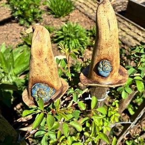 Könnte beinhalten: Zwei keramische Gartenornamente in Form von Hexenhüten. Die Hüte sind beige mit einem blauen Spiralmuster am Rand. Sie sind auf Holzpfosten montiert und von grünem Laub umgeben, was eine skurrile Gartendekoration ergibt.