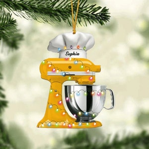 May include: A yellow stand mixer Christmas ornament with a chef's hat and the name "Sophia". The ornament is decorated with colorful string lights and a silver mixing bowl. A festive holiday decoration.
