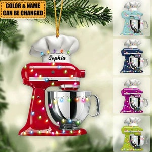 May include: A red kitchen mixer ornament with a chef's hat and the name "Sophia". The ornament is decorated with colorful string lights. Additional ornaments in blue, purple, and green are also shown.