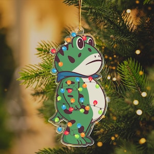 May include: A whimsical Christmas tree ornament featuring a cartoon frog with a grumpy expression. The green frog is adorned with colorful Christmas lights and a blue scarf. The ornament hangs from a gold string.