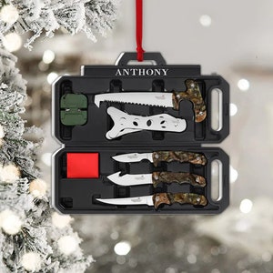 May include: Black toolbox-shaped ornament with the name "ANTHONY" in white. The ornament contains a saw, knives, and a sharpening tool. The handles of the knives have a camouflage pattern. A red ribbon is attached for hanging.