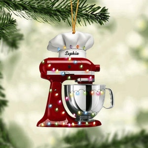 May include: A red stand mixer Christmas ornament with a white chef's hat and the name "Sophia". The ornament is decorated with colorful string lights. A silver mixing bowl is attached to the mixer.