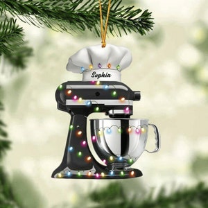 May include: A black stand mixer Christmas ornament with a chef's hat and the name "Sophia". The ornament is decorated with colorful string lights. The mixer has a silver bowl and is hanging from a gold string.