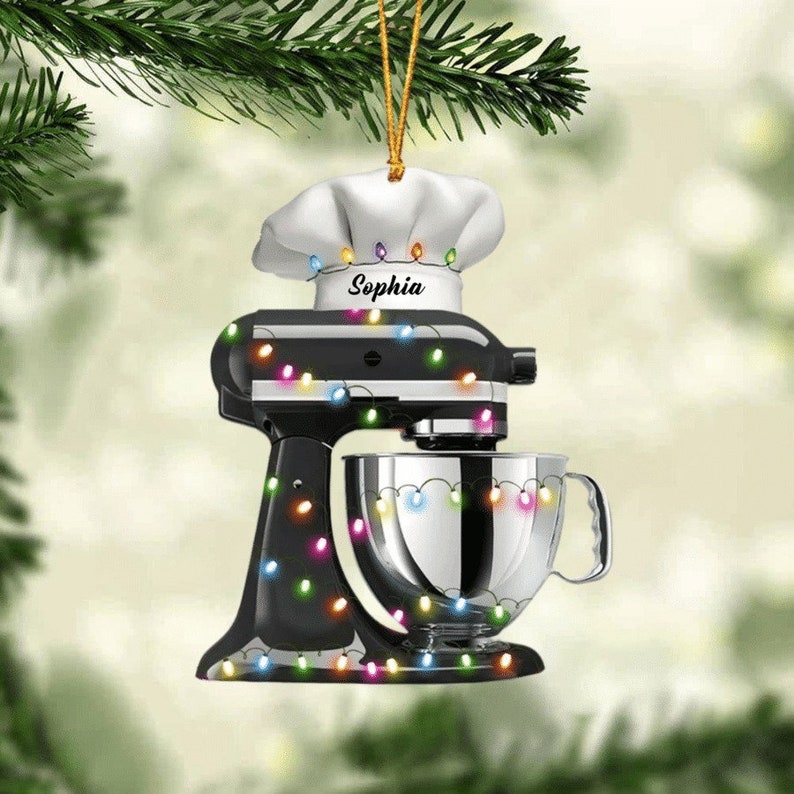May include: A black stand mixer Christmas ornament with a chef's hat and the name "Sophia". The ornament is decorated with colorful string lights. The mixer has a silver bowl and is hanging from a gold string.