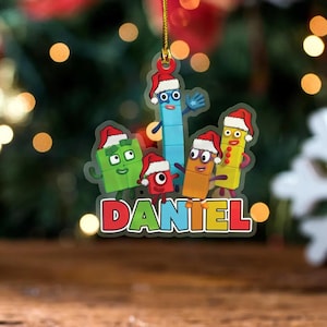May include: A flat, hanging Christmas ornament featuring a group of colourful, block-shaped cartoon characters wearing Santa hats. The characters are green, blue, red, orange, and yellow. The name DANIEL is printed in large, multicoloured letters.