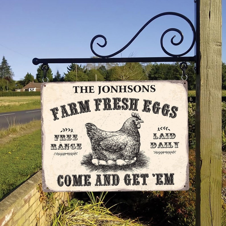 Op de afbeelding: Een metalen bord in vintage-stijl voor "Farm Fresh Eggs" met een zwart-wit illustratie van een kip en eieren. Het bord, met de tekst "The Johnsons", hangt aan een decoratieve zwarte metalen beugel en nodigt uit: "Come and Get 'Em".
