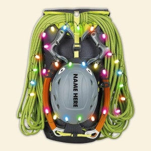 May include: A green climbing backpack with a helmet and climbing gear decorated with colorful Christmas lights. The helmet has the text "NAME HERE" printed on it.