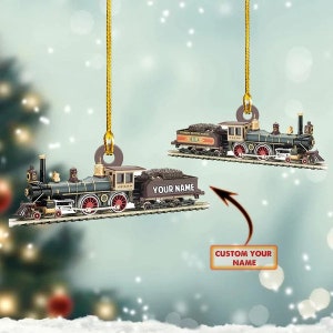 May include: A personalized Christmas ornament featuring a detailed model of a vintage steam locomotive train. The ornament is gold and features a black and red train with the words "YOUR NAME" on the side. The ornament is hanging from a gold string.
