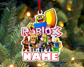 May include: A Roblox-themed Christmas ornament with a red ribbon for hanging. The ornament features the Roblox logo and various character figures. The word "NAME" is printed at the bottom. The ornament is set against a backdrop of a Christmas tree.