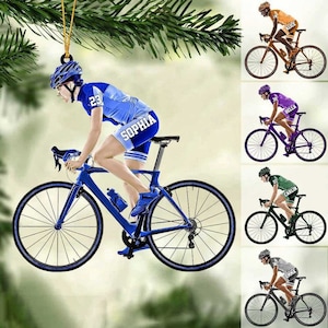 May include: A blue and white bicycle ornament with a cyclist wearing a blue jersey with the name "Sophia" on the back. The ornament is hanging from a green pine branch.