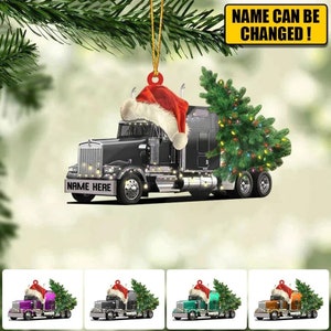 May include: A black semi-truck with a Christmas tree in the back and a Santa hat on the cab. The truck has lights on it and the text "NAME HERE" on the front bumper. The truck is a Christmas ornament.