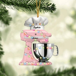 May include: A pink stand mixer Christmas ornament with a chef's hat and the name "Sophia". The ornament is decorated with colorful string lights and a silver mixing bowl. It hangs from a gold string.