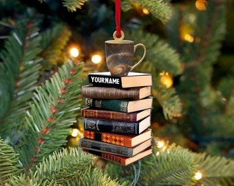 May include: A Christmas tree ornament featuring a stack of books topped with a coffee mug. The mug has a galaxy design. The top book has the text "YOURNAME". The books have various colors and textures, and the ornament hangs from a red ribbon.
