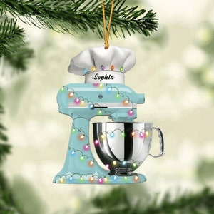 May include: A light blue stand mixer Christmas ornament with a chef's hat and the name "Sophia". The ornament is decorated with colorful string lights and a silver mixing bowl. A festive holiday decoration.