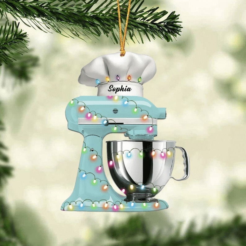 May include: A light blue stand mixer Christmas ornament with a chef's hat and the name "Sophia". The ornament is decorated with colorful string lights and a silver mixing bowl. A festive holiday decoration.