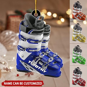 May include: A blue and white ski boot ornament with silver buckles and the name "Timothy" printed on the side. The ornament hangs from a gold string. Additional color variations of the ski boot are shown in orange, yellow, green, and red.