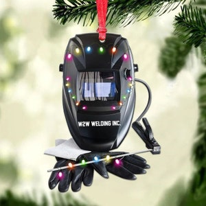 May include: A black welding helmet with a red ribbon and colorful Christmas lights around it. The helmet has the text "W2W WELDING INC." on it. The helmet is hanging from a Christmas tree.