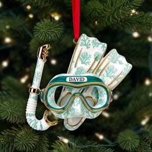 May include: A Christmas tree ornament shaped like a diving mask, snorkel, and fins. The ornament is white with teal coral and seashell designs, gold accents, and the name "DAVID" on the mask. A red ribbon is attached for hanging.