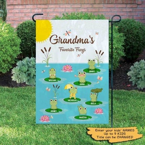 May include: A personalized garden flag with a blue background and a sun in the top left corner. The text "Grandma's Favorite Frogs" is in the center of the flag. There are nine frogs with names written below them, including William, Anna, Jimmy, Nicholas, Bella, Henry, and Martha.