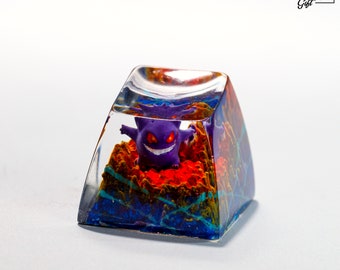 May include: A clear resin keycap with a purple and red Gengar Pokemon figure inside. The resin is filled with blue, green, and orange swirls.