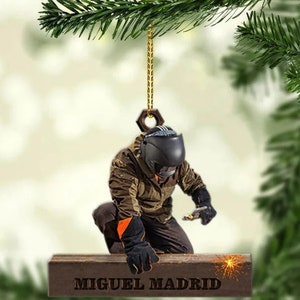 May include: A personalized Christmas ornament featuring a welder wearing a welding mask and welding a piece of metal. The ornament is brown and has the name "Miguel Madrid" engraved on it.