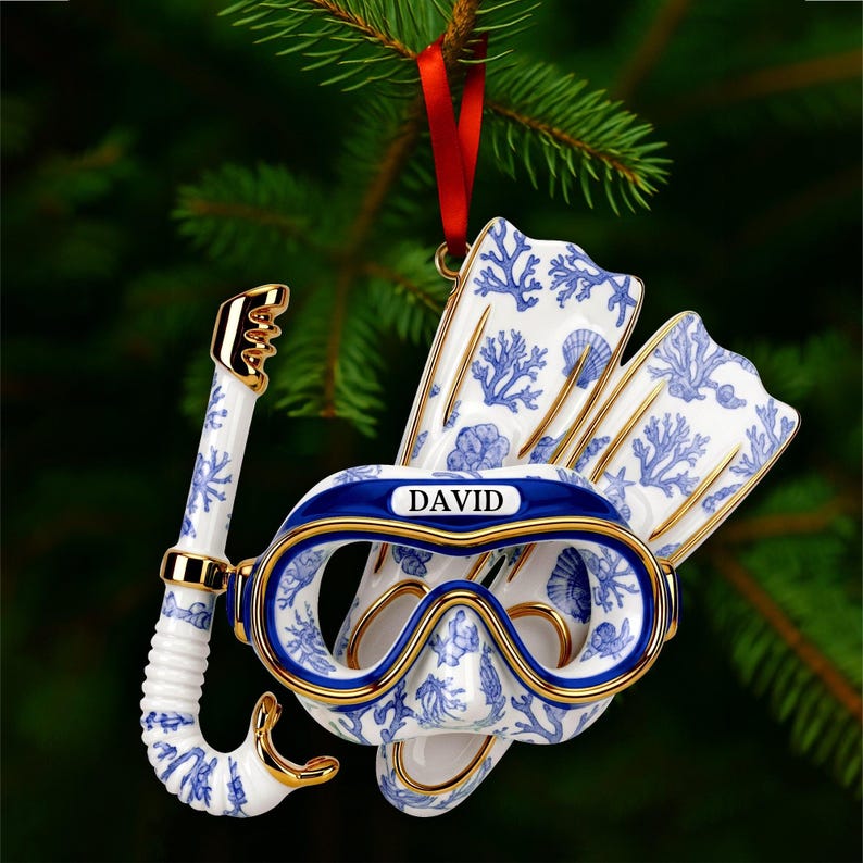 May include: A ceramic Christmas ornament shaped like a diving mask, snorkel, and fins. The ornament is white with blue coral and seashell patterns, gold trim, and the name "DAVID" on the mask. A red ribbon is attached for hanging.