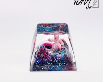 May include: A clear resin keycap with a pink Pokemon figurine inside. The resin is filled with blue, pink, and silver glitter, creating a galaxy-like effect. The keycap is labeled "HAVI Gift".