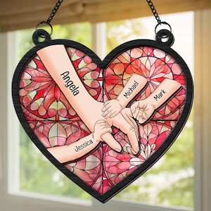 May include: A stained glass heart-shaped ornament with a pink and red design. Four hands are intertwined, with the names "Angela", "Jessica", "Michael", and "Mark" written on the arms.