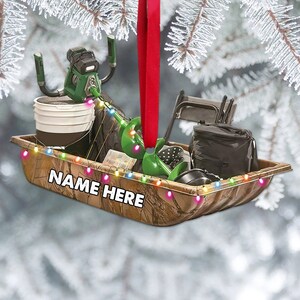 May include: A Christmas ornament shaped like a camo sled filled with ice fishing gear, including a drill, bucket, and chairs. The sled has a string of colorful lights and the text "NAME HERE" on the side.