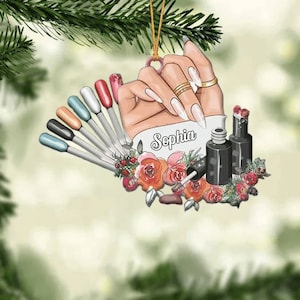 May include: A Christmas ornament featuring a hand with long, white nails holding a fan of colorful nail polish bottles. The ornament is decorated with red roses and the name "Sophia" is written on the ornament.